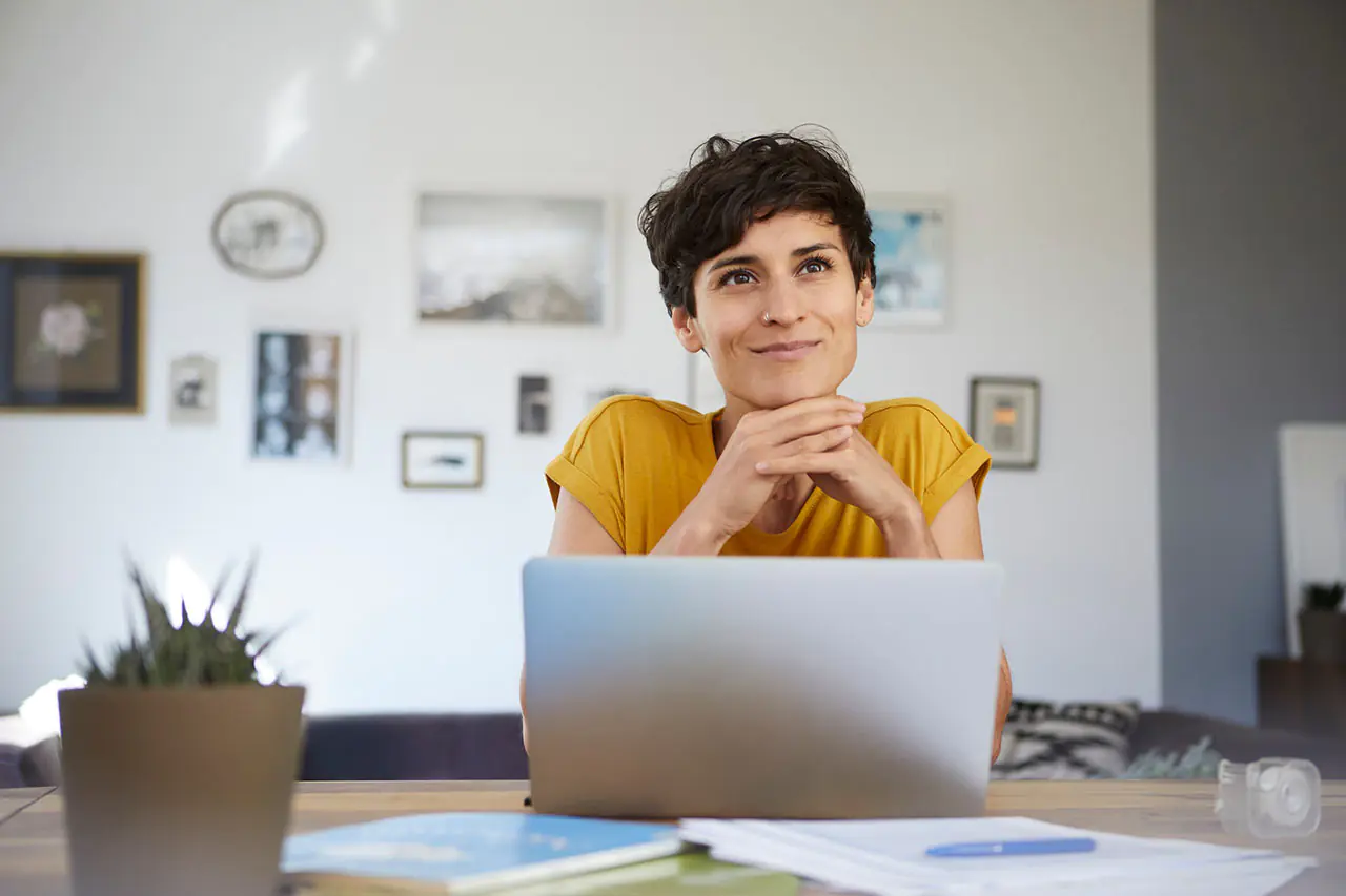 Person mit gelbem T-Shirt sitzt an einem Tisch vor einem Laptop, im Hintergrund mehrere gerahmte Bilder an der Wand.
